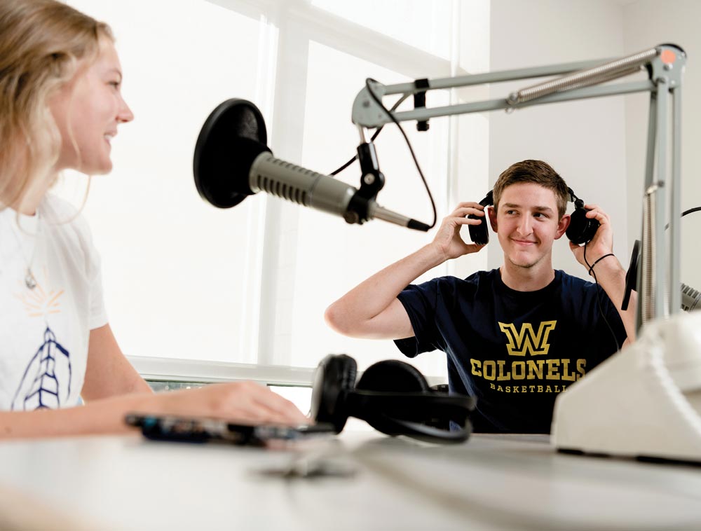 Two students talking to eachother in front of recording microphone with one also wearing a headset