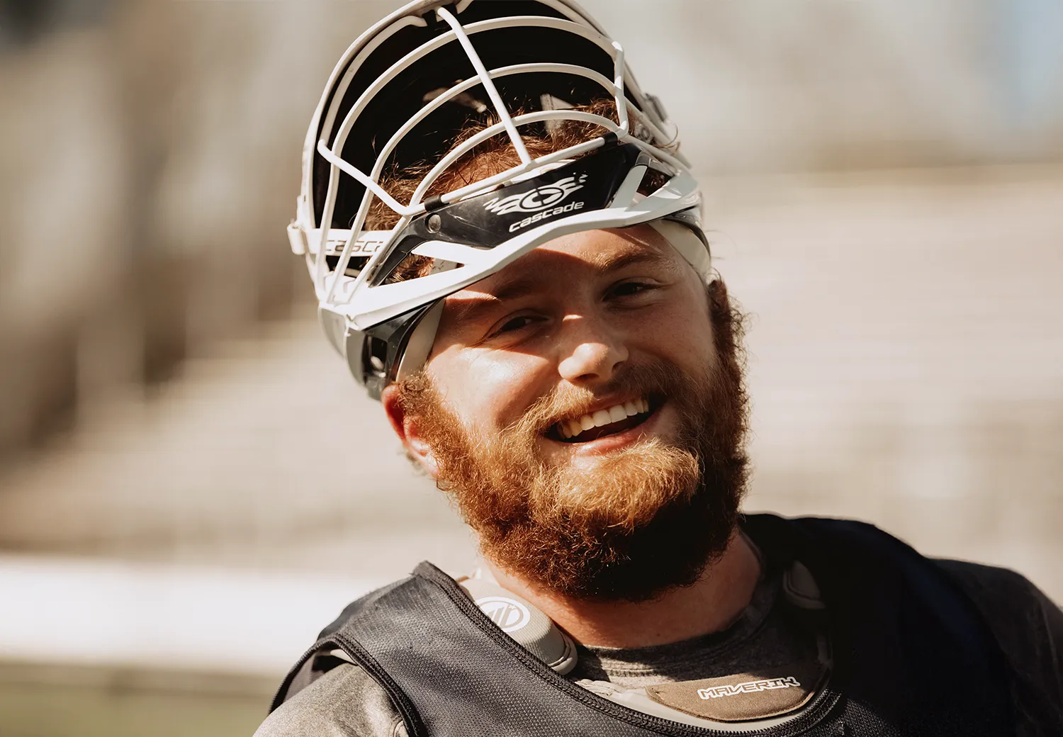 Male lacrosse player smiling with his helmet tilted back on his head, wearing protective gear on a sunny day.