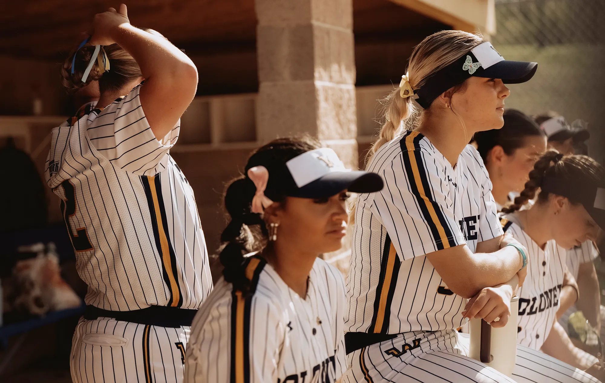 Softball players in striped uniforms sitting in the dugout, one adjusting her hair while others watch the game.