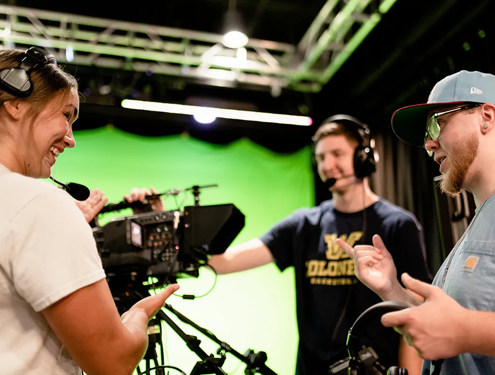 Three students working together in a television production studio. One operates a professional camera while the others, wearing headsets, smile and discuss the setup in front of a green screen.