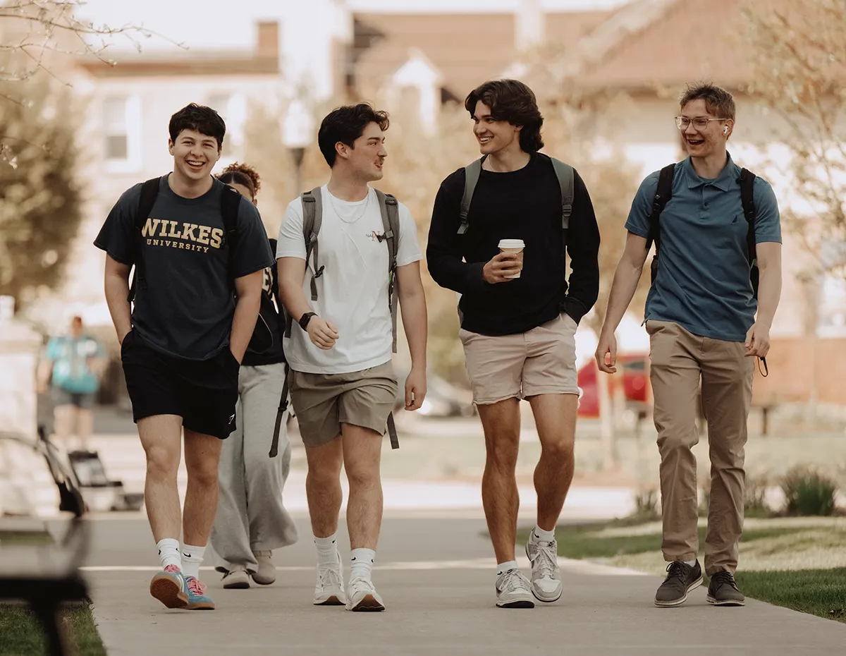 Four college students walking together on campus, smiling and talking. One wears a “Wilkes University” T-shirt, another carries a coffee cup, and all have backpacks.